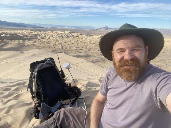 A bearded man in a green hat sits next to a ham radio and a backpack in the foreground, the background is a sea of sand dunes.