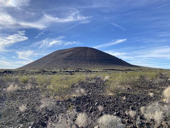 A volcanic cinder cone of red and black lava sits about 1 mile from the viewer rising from the Mojave desert floor