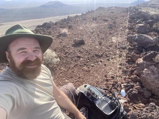 A bearded man in a green hat, sits next to a ham radio, and an antenna in a reddish volcanic landscape at the edge of a large volcanic cinder cone.