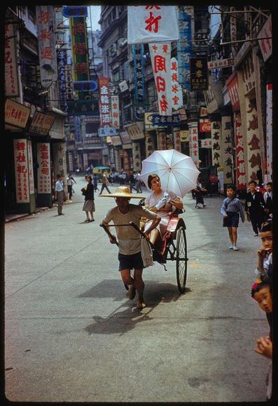 A man pulling a woman in an old-fashioned bicycle rickshaw down a street, lined with Chinese businesses and advertisements. The scene is set during the day, under sunlight that casts shadows on the ground. People are walking by or standing along the streetside; some look directly at the camera while others engage in various activities such as talking on mobile phones.