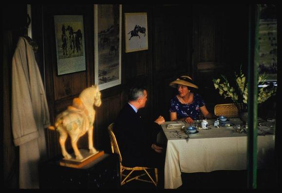 The image is a color transparency photograph taken in 1959. It features an intimate indoor scene with two individuals seated at a table, engaged in conversation over tea and pastries. The setting appears to be a sophisticated dining area adorned with elegant artworks depicting equestrian scenes on the walls behind them.

On one side of the room stands a porcelain horse statue, while another person's coat hangs casually near it. A woman is wearing a wide-brimmed hat, indicative of fashion trends or perhaps an outdoor excursion earlier in their day; she has her hair styled back and wears minimal jewelry with large earrings visible as they sit at the table.

The man appears to be dressed formally for dinner or tea service during this era but leans slightly forward into his companion's space. The woman seems more relaxed, leaning on one hand while conversing over a teacup placed in front of her on an ornate porcelain saucer with matching cup and bowl set beneath it; there are other similar sets across the table as well.

The overall ambiance suggests that this photograph captures a moment from social life within Hong Kong's English colony during its colonial history. The image is part of "Sightseeing in Hong Kong" by Sports Illustrated, which was published on July 1959 and includes notable figures like Toni Frissell (photographer) known for her work with baseball subjects suc [...]