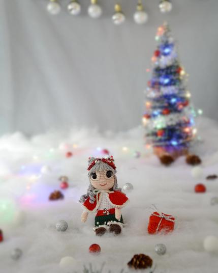 Poupée au crochet représentant une lutine de Noël assise dans la neige. Elle porte une tenue rouge et blanche, et des lunettes rondes. Elle a une chevelure grise avec 2 petits chignons sur la tête, décorés d'une guirlande rouge et d'un grelot. Un petit cadeau est posé au sol à côté d'elle, avec un sapin de Noël lumineux flouté en arrière-plan.