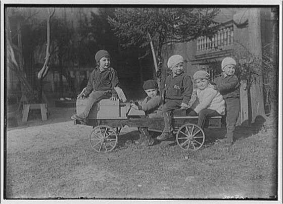 The image depicts a group of four children posing for the camera with vintage wagons and boxes. These wagons appear to be made from wood, featuring simple wheels and handles on both ends. One child is standing beside his wagon, while another sits atop it holding onto its boxy back. The third child is seated within one of these wagons, which seems filled with items like a suitcase or crate. Finally, the fourth child stands next to an empty wagon.

The setting appears to be outdoors during daytime hours in what could possibly be a backyard given hints at residential structures and vegetation in the background. Notably, all children are wearing hats or headgear that suggests it may be cold outside as they're dressed warmly in coats and trousers appropriate for winter attire of early 20th-century America.

The photograph is rendered in black and white due to its age, likely taken during a time when color photography was not yet prevalent. It captures the innocence and charm typical of children from that era while also serving as an artifact reflecting social norms, fashion trends, and possibly socioeconomic status based on their attire and surroundings.
