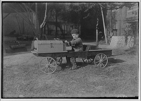 The image depicts a young child seated on an old-fashioned wagon, with one foot placed on the ground and holding onto what appears to be a handle or steering mechanism. The wooden cart has two wheels at its front end and is positioned outdoors in a grassy area surrounded by trees and some makeshift structures that could suggest it's near a fairground or carnival setting.

In the background, there are various objects such as tents or booths with white canvas canopies visible behind the wagon, along with other indistinct items like a small boat on water. The photograph is in black-and-white, which gives it an aged appearance consistent with early 20th-century photography techniques.

The overall atmosphere of the image conveys a sense of nostalgia and simplicity associated with childhood play during that era.