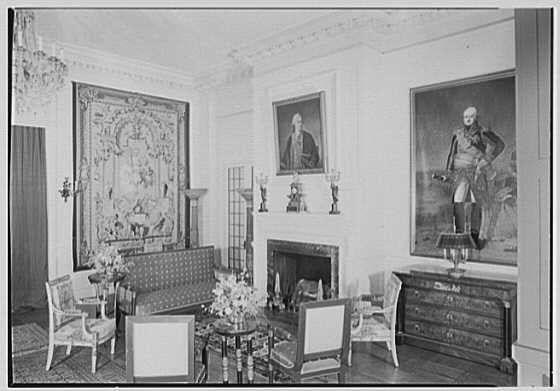 The image depicts a formal living room with antique furnishings and decor, captured as an archival photograph. The large space is illuminated by natural light entering through unseen windows.

In the center of the room stands a fireplace framed by two life-size portraits; above are more paintings hung on white walls. A grand tapestry embellishes one wall. Furniture includes upholstered chairs, with some bearing intricate patterns and designs indicative of an older style. An ornate wooden sideboard is visible to the right.

The photograph's monochromatic tone suggests it was taken in a bygone era, likely around 1945 as indicated by the caption mentioning "Feb. 17." The source is credited to Gottschol-Schleisner Inc., and details such as negative size are included for cataloging purposes.

The ambiance exudes elegance with its classical elements and historical decorations, offering a glimpse into an era of formal design sensibilities in interior decoration.