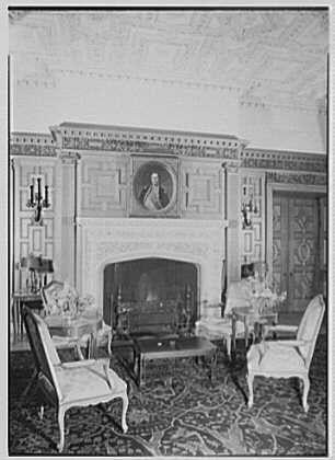 The image shows a historical black and white photograph of an elegantly furnished living room with ornate details. A large portrait hangs above the fireplace, which is adorned with decorative mantels on either side. The walls are decorated with intricate patterns, possibly frescoes or painted plasterwork.

Two chairs with classic design elements flank each side of the fireplace, accompanied by a couple of lamps and vases that add to the room's vintage charm. A carpet with a floral pattern covers the floor, complementing the overall antique aesthetic of the space. The absence of any modern appliances suggests this photo captures an older period or represents a preserved historical setting.

The caption indicates the photograph is from Gottscho-Schleisner Inc., likely referencing a company involved in photography or artistry during that era, and notes it as part of the French Embassy collection at Washington D.C. Library I dated February 17th, 1945. The image has one negative with dimensions of approximately 5x7 inches.