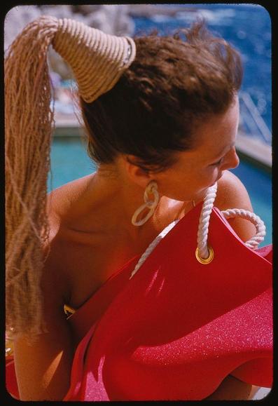 A woman wearing a red swimsuit, sitting near the poolside and embracing her bright pink handbag. She is wearing earrings with seashells on it and has an updo style ponytail which seems to be made of rope-like fabric that extends down her back.

Toni Frissell was famous for photographing celebrities during the 1940s and '50s, capturing iconic images like this one. This photo is a part of Sports Illustrated's feature on Dior models from August, 1954; Eden Roc hotel in Miami Beach serves as an exclusive backdrop.

Christian Dior was a renowned fashion designer known for his "New Look" collection that revolutionized women’s fashion post-World War II era. The image shows Lucky posing elegantly with her bright red handbag and accessories made of seashells, which is fitting given the association with high-end brands like Christian Dior.

In conclusion, this photograph captures a moment from Sports Illustrated's August 1954 edition featuring model "Lucky" showcasing fashion trends associated with renowned designer Christian Dior. The Eden Roc hotel in Miami Beach provides an exotic and luxurious atmosphere typical of upscale publications such as Sports Illustrated during that period.