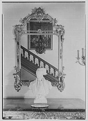 This black and white photograph shows an elegant console table with a large decorative mirror mounted on it. The mirror reflects the image of a staircase, which appears to be part of a grand interior space adorned with classical architecture.
Atop the console table is a bust sculpture or figurine, seemingly made of marble or another stone material. In the background, partially obscured by the reflection in the mirror and not fully visible due to perspective, there seems to be an artwork depicting figures that could represent historical or allegorical scenes.
The overall setting suggests a formal space with classical design elements typical for diplomatic or high-society venues during the early 20th century. The photograph bears metadata indicating it was taken on February 17, 1945, at the French Embassy in Washington D.C., specifically within an entrance hall that features this console table and mirror as part of its decorative scheme.