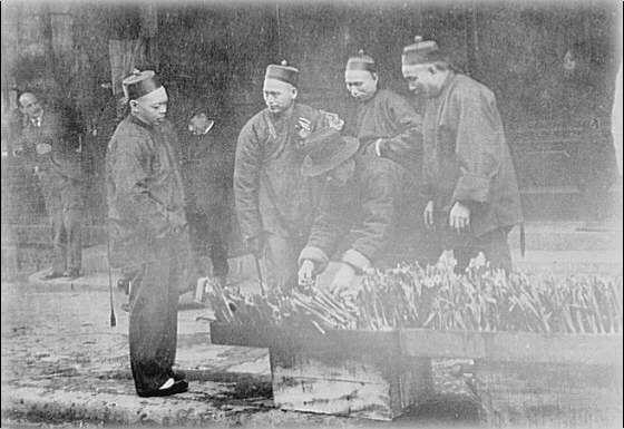The image is a black and white photograph showing four men, likely vendors from the late 19th or early 20th century. They are standing around a large tray filled with what appears to be sticks of incense or perhaps wooden items for sale in an outdoor market setting.

Each man wears traditional East Asian attire, including robes and conical hats known as "bailin" caps typically worn by Chinese men during that era. The first man on the left stands upright with a cane in hand while observing his surroundings. His posture suggests contemplation or mild interest.

The second and third individuals are actively engaging with each other over the tray of items, possibly discussing their sale price or quality. Their expressions seem to convey concentration and collaboration as they interact closely.

The fourth man on the right is partially turned towards the others, suggesting he may be more observant than directly participating in the conversation at that moment.

In the background, another person can be seen walking away from the group, further establishing a bustling street scene. The image captures an authentic glimpse into daily life and commerce within Chinatown during Genthe's photography era.