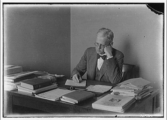A black and white photograph of a man sitting at his desk, writing on some papers with several books around him. The image is described as "Portrait photographs" in Portrait of Mr. True seated at his desk II ca. 1920-ca. 1950. Horydczak, Theodor, approximately 1890-1971 and has a negative size mentioned as 6x4 inches.