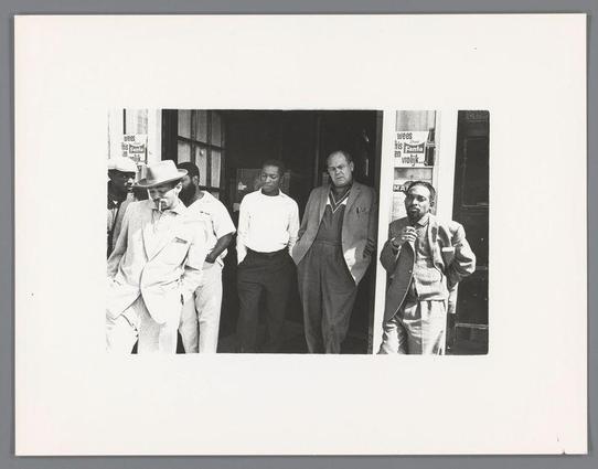 The image shows a group of five men standing in front of what appears to be the entrance of an establishment. The setting is urban, with signs and storefronts visible through glass windows behind them. From left to right: One man wears sunglasses atop his head, another has on a light-colored suit jacket over casual clothing, a third stands casually dressed with hands near his chest, followed by a fourth in business attire holding a cup of coffee or tea, while the last is clad in a dark coat and appears contemplative. The black-and-white photograph captures them mid-stride as they exit through double doors into daylight. Posters on the window contain text related to beverages such as "wees fris Fanta" which suggests commercial activity nearby. There's no explicit indication of their occupations or intentions, but it seems like a candid moment captured in public space during an earlier era based on clothing and photographic style.