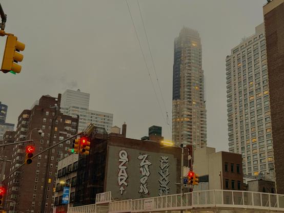 A grey, gloomy day in Manhattan. Red traffic lights are lit up. Yellowy orange lights from rooms in apartment buildings are also on display. Otherwise the sky is grey and overcast.