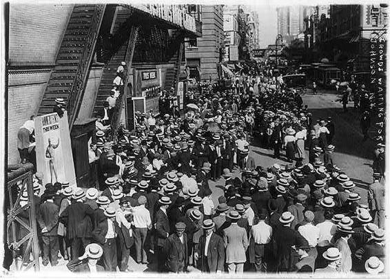 The image is a black-and-white photograph depicting a large crowd gathered in an urban setting. The scene appears to be from the early 20th century, judging by the clothing styles and architectural features. The crowd is densely packed, with individuals wearing suits, hats, and other formal attire typical of the era. Some people are standing on the steps leading up to a building, while others are standing on the street, suggesting a public event or gathering.

The building in the background has a sign that reads "Hirsch's," indicating the name of a business or establishment. The architecture of the building and the surrounding structures suggests a city environment, possibly a commercial or business district. The street is lined with other buildings, and the overall atmosphere conveys a sense of activity and public interest. The image has a vintage quality, with a slightly grainy texture, which is characteristic of early photographic prints.