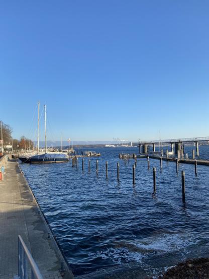 Sporthafen an der südlichen Kiellinie in der Kieler Förde. Fast alle Dalben sind leer, lediglich zwei Segelboote liegen noch im Wasser. Das leicht wellige Wasser der Ostsee schäumt an der Kaimauer, im Hintergrund ist ein DFDS-Fährschiff am Ostuferhafen neben dem Küstengaskraftwerk zu sehen. Der Himmel ist strahlend blau, am Horizont türmt sich ein Wolkenberg auf, der auf den ersten Blick eher nach einer Gebirgskette aussieht.