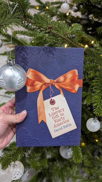 Holding the book "The Literary Gift in Early America" by David Faflik in front of a large Christmas tree, decorated with silver-white ornaments