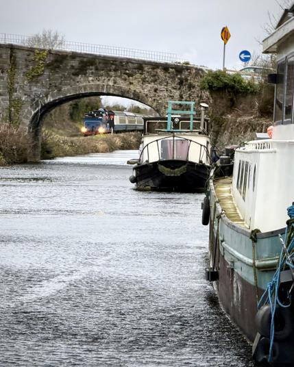 A telephoto view looking through the arch of a stone bridge over a canal at a blue steam train approaching in the distance. The edges of canal barges moored in front of the bridge frame the right of the scene.