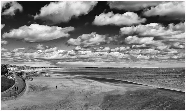 A black and white image of a beach scene featuring a wide sandy shoreline, calm waters, and a cloudy sky. A few people can be seen walking along a path near the beach, with a distant coastline and structures visible in the background.