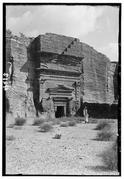 This black and white image depicts an ancient rock-cut tomb located in a barren landscape. The structure appears to be carved into the side of a cliff, showcasing architectural elements such as pilasters (flattened columns) and a pedimented doorway that suggest classical influence. Above this entrance is what looks like Nabatean capitals - rounded finials at the end of columns or architraves in ancient Greek architecture.
In front of the tomb stands an individual wearing traditional Middle Eastern attire, including a long robe with wide sleeves and head covering, which gives context to the era being represented. The terrain surrounding this structure is arid, featuring sparse vegetation such as small bushes and dry patches indicative of desert conditions.
The photograph bears a handwritten notation in the upper left corner that reads "C-13" alongside an illegible symbol or signature below it, possibly serving as a cataloging identifier for Archival purposes.