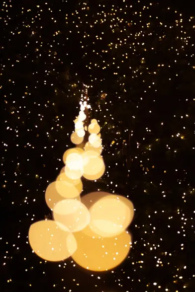 Mittig Bokehbubbles und -katzenaugen in warmtonig im Verlauf nach hinten auf die Sternenhimmelartige Beleuchtung des großen Weihnachtsbaumes zu. Hintergrund dunkel mit hellen Lichtpünktchen.