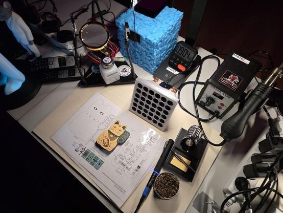 A cluttered workspace featuring a variety of tools and items: a magnifying glass, soldering iron, electronic components, a sketch with a cat design, and multiple gadgets including old mobile phones. A blue 3D-printed object and a small