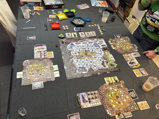 A tabletop game setup is displayed, featuring a large, intricate game board with various hexagonal tiles and player areas. Game pieces, cards, and tokens are scattered across a dark tablecloth, alongside cups and snacks. One player is visible, wearing a christmas like sweat shirt