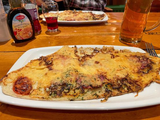 Auf einem hölzernen Restauranttisch stehen zwei rechteckige Teller mit Pfannkuchen, jeweils in Form der holländischen Insel Texel. Sie sind mit Käse überbacken und verbergen verschiedene Produkte, die von der Insel stammen. Neben einem Glas Limonade und einem Krug Bier stehen noch Puderzucker und Zuckersirup auf dem Tisch.