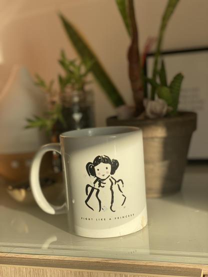 White coffee cup sunlit on a side table with plants in the background. Stenciled drawing of a woman with buns on each side of her hair and text “fight like a princess” underneath.