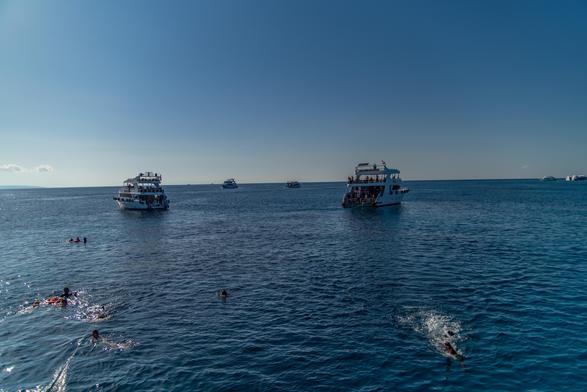 Obraz przedstawia kilka łodzi na spokojnym niebieskim oceanie, z kilkoma ludźmi pływającymi w pobliżu. Niebo jest czyste i słoneczne, tworząc ciepłą, spokojną atmosferę.