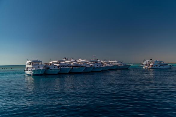 Kolejka białych łodzi zakotwiczonych w spokojnych, błękitnych wodach pod czystym niebem, z niektórymi łodziami widocznymi w tle.