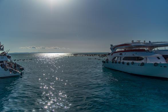 Spokojny pejzaż morski z dwiema białymi łodziami zakotwiczonymi w spokojnej, lśniącej wodzie. W tle wiele osób cieszy się wodą, a łagodne fale odbijają światło słoneczne. Niebo jest czyste z kilkoma chmurami, tworząc spokojną atmosferę.