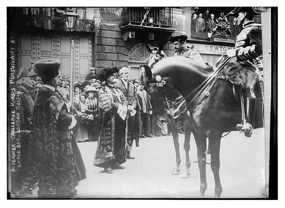 This is a historical black-and-white photograph depicting a formal event in London, as indicated by the text on the image. The scene shows a group of people gathered in what appears to be a public space, possibly a street or a square. In the foreground, a man on horseback is prominently featured. He is dressed in a military-style uniform, complete with a hat and a sword, suggesting he may be a soldier or a ceremonial guard. The horse is adorned with a saddle and bridle, and the rider holds a reins and a flag, indicating a ceremonial role.
Behind the horse, a group of people is standing, dressed in formal attire typical of the early 20th century. The women are wearing long dresses with intricate patterns, while the men are in suits and top hats. The crowd appears to be observing the event, with some individuals looking towards the horse and rider, possibly indicating a moment of attention or anticipation. The background features a building with a sign that reads "Newton," and other architectural details, such as a large door and decorative elements, are visible. The overall atmosphere of the image conveys a sense of formality and importance, likely related to a significant public event or ceremony.