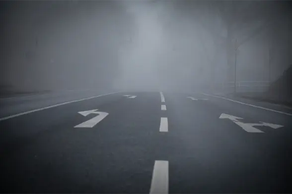 A photograph of an inner-city street in the fog. The photographer was standing in the middle of the street. On the asphalt, two turning lanes can be seen, marked with white lines and arrows. The end of the street is not visible.