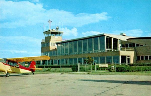 A color postcard view shows the passenger terminal and control tower at St. Joseph County Airport, also known as Bendix Field, in South Bend, Indiana. The main building is a low, mid-20th-century terminal constructed of light-colored brick with long horizontal bands of large glass windows on the upper level. A square control tower rises above the terminal, topped with antennas and aviation equipment. In the foreground, on the concrete apron, a small single-engine propeller airplane is parked facing left; it is painted yellow with red accents on the tail and fuselage. A chain-link fence separates the aircraft area from neatly trimmed grass and shrubs around the terminal. The sky above is bright blue with scattered white clouds, indicating clear weather conditions.