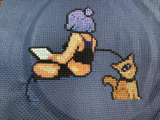 Broderie au point de croix d’une femme de trois quart dos, sur un bean bag, avec un laptop. Au premier plan, un chat mutant avec un grand œil unique, vous regarde avec un petit sourire.