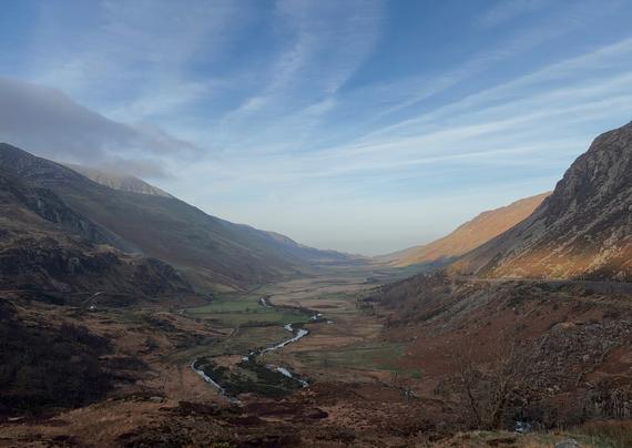 A wide, U-shaped glacial valley in North Wales under a blue sky with wispy clouds. A small river meanders through the flat, grassy valley floor between steep, rugged mountains. The mountain slopes are covered in brown heather and patches of green, with sunlight hitting the right-hand ridge.