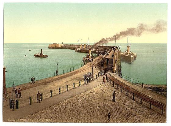 The image depicts a vintage scene at Dover Admiralty Pier in England, dating approximately between the late 19th and early 20th centuries. A prominent feature is a large ramp leading from the land down to the water's edge where several ships are moored or docked, with one ship emitting black smoke indicating it may be powered by coal steam engines common during that era.
The pier itself extends into calm waters under an overcast sky with hints of blue peeking through. Alongside the paved walkway at the top level is a railing and street lamps, suggesting pedestrian pathways for visitors or workers. Several individuals dressed in period-appropriate attire are scattered across the scene: some walking along the promenade, others standing near lampposts.
The image captures an industrial port bustling with maritime activity against the backdrop of British seaside architecture. The overall atmosphere is one of historical significance and a glimpse into life during that time at Dover's Admiralty Pier, likely serving as a strategic entry point for naval vessels or merchant ships navigating through English waters.