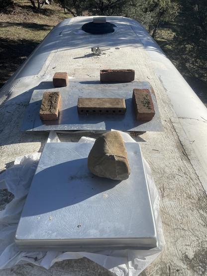 Roof of a vintage trailer with white trash backs secured around a skylight in the foreground covered by an aluminum baking pan weighted down with a rock.

Fan/skylight behind that.  Also covered with white trash bags (not visible) then a sheet of aluminum weighted by bricks.

Another skylight in the distance with the same strategy covered by a trashcan lid weighted down by a paver.