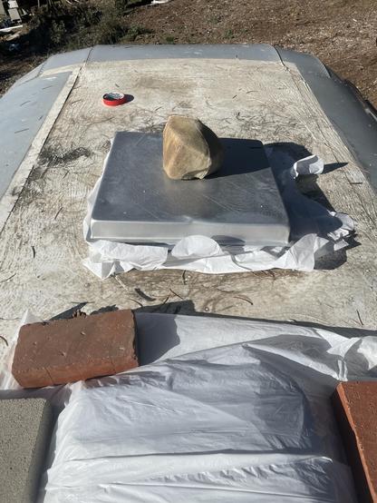 Roof of a vintage trailer with white trash backs secured around a skylight in the foreground secured by bungies covered by an aluminum baking pan weighted down with a rock.

Fan/skylight in front of that.  Also covered with white trash bags weighted by bricks.