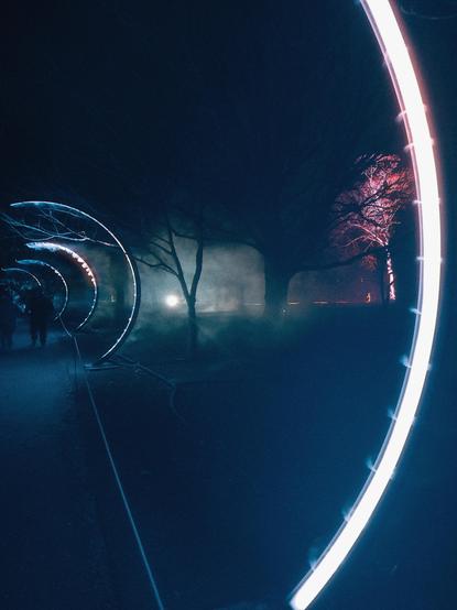 A dimly lit pathway features illuminated circular installations alongside trees. Fog creates an atmospheric effect, and a few silhouettes of people can be seen walking through the scene.