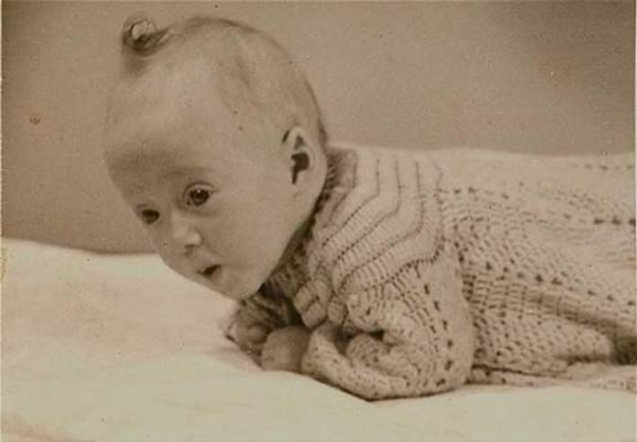 A vintage photo of a baby in a knitted sweater, lying on their stomach and looking to the side with a curious expression.