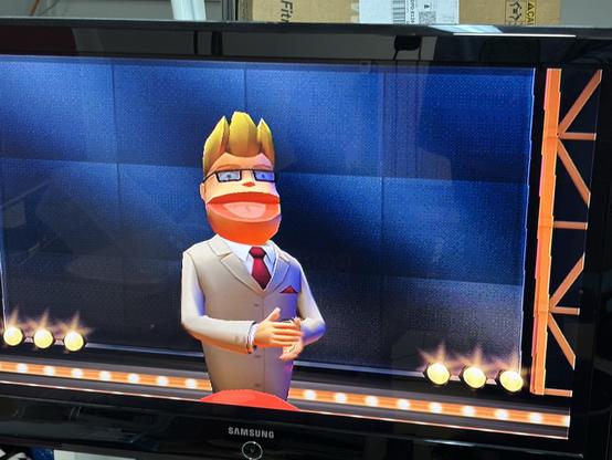 Das Bild zeigt eine animierte Figur aus dem PlayStation 2-Spiel „Buzz“ mit blonden Haaren und Brille, gekleidet in einen leichten Anzug und eine rote Krawatte, die auf einer Bühne mit Lichtern in einem Fernsehstudio steht. Der Hintergrund zeigt eine blaue Wand mit einem Scheinwerfereffekt.