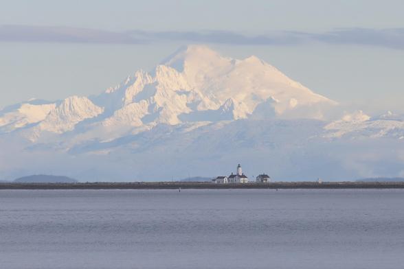 A massive snow-covered volcano looming in the distance with a cute little lighthouse in the foreground.
