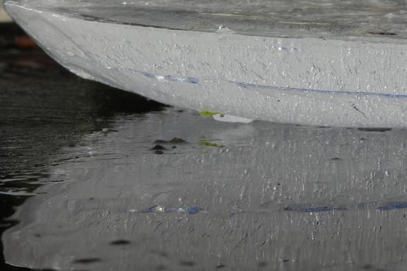 A round piece of ice, resting on top of a black shard of glass, is illuminated from the side by the sun. Countless tiny bubbles rise very slowly within the ice, leaving traces of their movement. The bubbles reflect the shard and the light. Some bubbles shimmer with spectral colours due to refraction. A tiny, light green piece of moss, frozen to the lower part of the piece of ice, and its reflection can be seen on the left of the image. The ice piece is reflected, partially distorted, in the black glass that is covered with a thin film of ice and water. A narrow ridge at the edge of the ice reflects the blue of the sky.