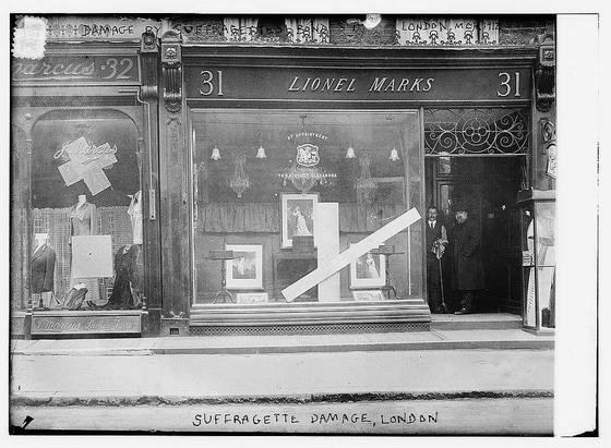 The image is a historical black-and-white photograph of a storefront in London, dated 1912. The shop is labeled with the name "Lionel Marks" and has the address "31" above the entrance. The storefront has a large display window showcasing mannequins dressed in formal attire, likely for women, with some clothing items laid out on the floor. The display window is marked with a large "X" and the words "Suffragette Damage" written on it, indicating that the shop was vandalized by suffragettes, a group advocating for women's suffrage. The shop's interior appears to have ornate decorations, including a chandelier and intricate metalwork on the door frame. Two men are standing in the doorway, seemingly observing the damage. The overall scene reflects the social and political tensions of the early 20th century, specifically related to the women's suffrage movement.