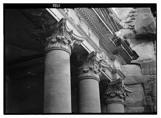 The image depicts a historical architectural structure, showcasing three ornate columns with intricate capitals. These columns appear to be part of an ancient building or monument, possibly situated in Petra (Wadi Musa), as indicated by the context provided.
Each column features elaborate decorative work at its top, including what appears to be acanthus leaves and other classical motifs that suggest a Greco-Roman influence on Roman architecture. The capitals are richly detailed with scrollwork, volutes, and leafy patterns, indicating skilled craftsmanship. There is visible wear and tear on the columns' surface, hinting at their age.
The photograph seems to be taken from an angle slightly below eye level, emphasizing the height of the pillars and providing a clear view of the architectural details. The black-and-white nature of the image suggests it may have been captured during or shortly after its construction in approximately 1920-1933. This photo is part of a collection by American Colony Photo Department located in Jerusalem, as referenced on the negative.
The overall composition focuses on highlighting the beauty and craftsmanship of these ancient columns amidst their aged stone surroundings, capturing an essence of historical significance and architectural grandeur from that era.