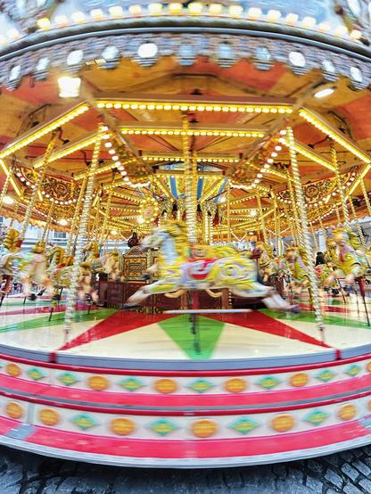 A brightly-coloured and brightly lit merry-go-round. The wooden, white-and-gold horses gallop clockwise (with motion blur), unencumbered by riders. It was 10 am and the carousel had only just opened for business. Stall Street, Bath, UK. Photo by Gearóid Burke.