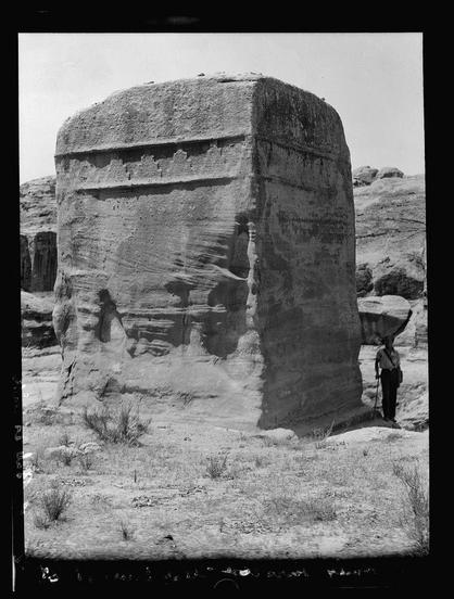 The image displays a large, ancient stone structure with visible signs of weathering and erosion. It appears to be made from sandstone or similar material, showing various striations and lines that suggest it has been exposed to the elements for an extended period.
In front of this monolithic edifice stands a person who seems small in comparison due to the imposing size of the structure. The individual is dressed casually with what looks like long pants and boots, likely suggesting outdoor or archeological activity. They are extending one arm upwards towards the structure's top edge as if pointing out features on it.
The surrounding environment includes dry ground with sparse vegetation indicative of a desert-like terrain, which might suggest this photo was taken in an area known for such rock formations like Petra (Wadi Musa), as noted at the bottom. The sky is clear and blue, implying fair weather conditions during when the photograph was captured.
Overall, it presents a scene likely from a historical or archaeological exploration site with natural features characteristic of ancient civilizations that built monumental structures in arid regions for various purposes such as religious worship, memorials, or territorial markers.