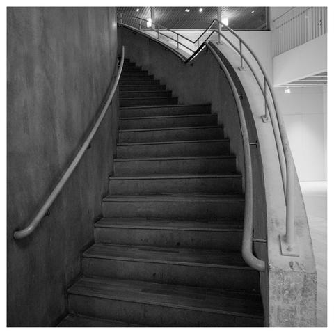 En fyrkantig svartvit bild på en trappa uppåt som viker kurvar sig åt vänster och försvinner. Ett räcke på var sida och ett extra högre räcke på höger sida. Till vänster en vägg. Några lampor lyser genom fönster på övre våningen.