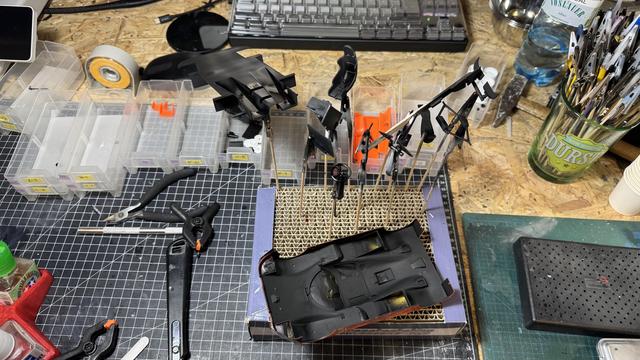 An overhead view of a modelling workstation with many small painted model parts held on skewers in a corrugated cardboard stand. A black painted inner body tub sits in a box on the right. Tools, storage bins and a keyboard are visible in the background.