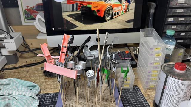 Close-up of many small painted model pieces on skewers drying in a cardboard holder in front of a computer monitor. Painted wheels, body panels and green aero parts are visible; the monitor displays a bright orange race car photo.